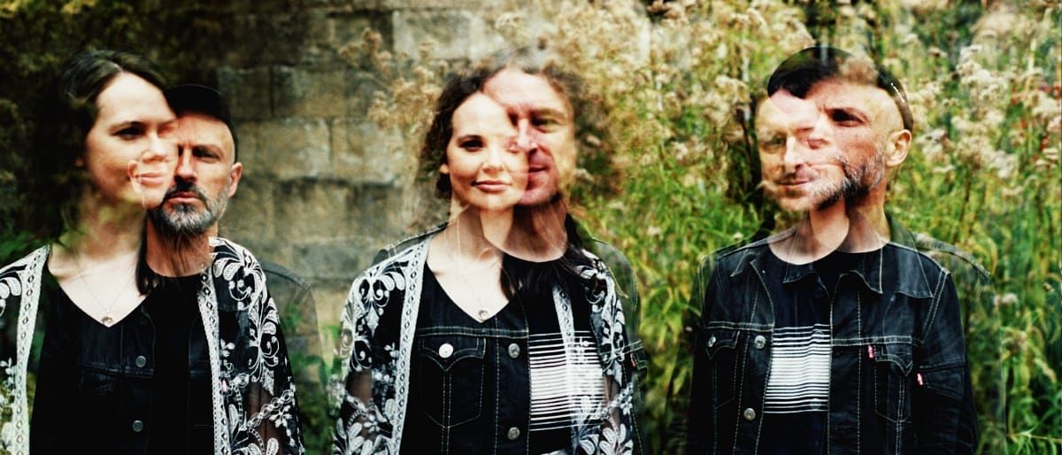 A double-exposure outdoor photo of Golden Samphire Band, three members standing before wild grasses and a stone wall, each face layered with a ghosted second image.