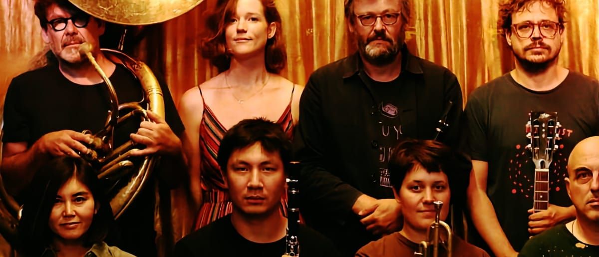Eight members of The Notwist cluster together before a gold curtain, several holding instruments including a sousaphone, guitar, clarinet, and trumpet.