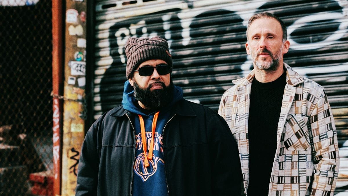 Dälek stand in front of a graffiti-covered metal shutter, one in a knit cap and sunglasses with a Knicks hoodie, the other in a patterned coat. Photo by Jonny Scala.
