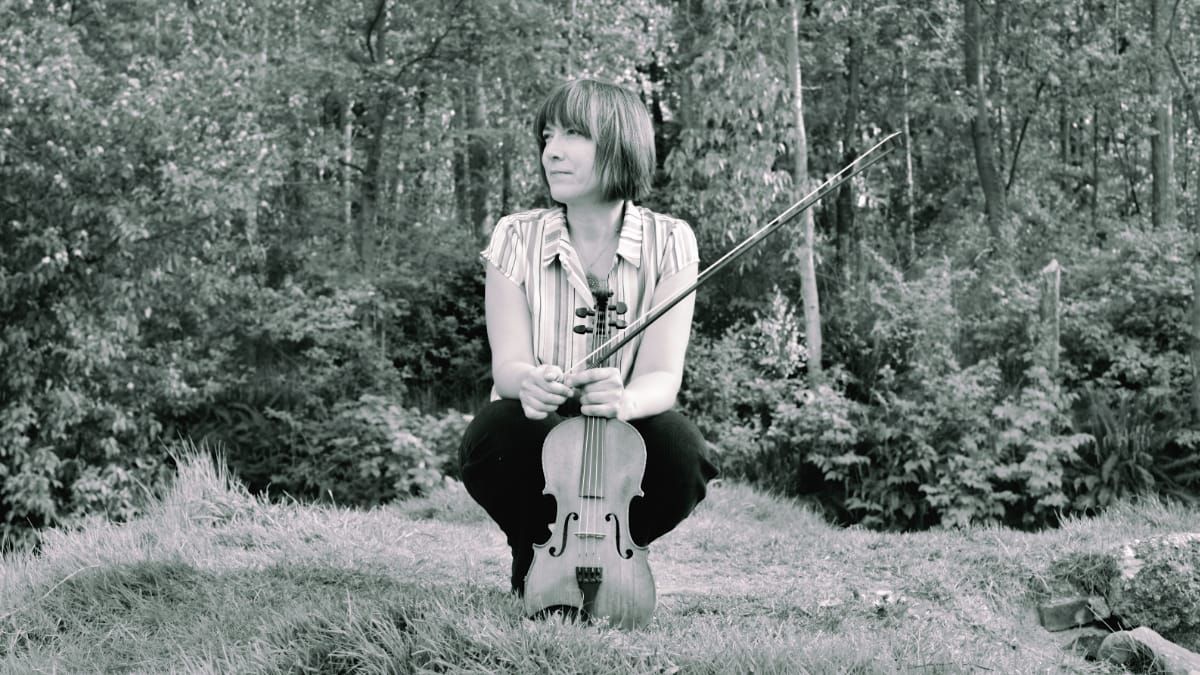 Meredith Bates crouches on grass before a wooded backdrop, holding a violin and bow, gazing to one side.