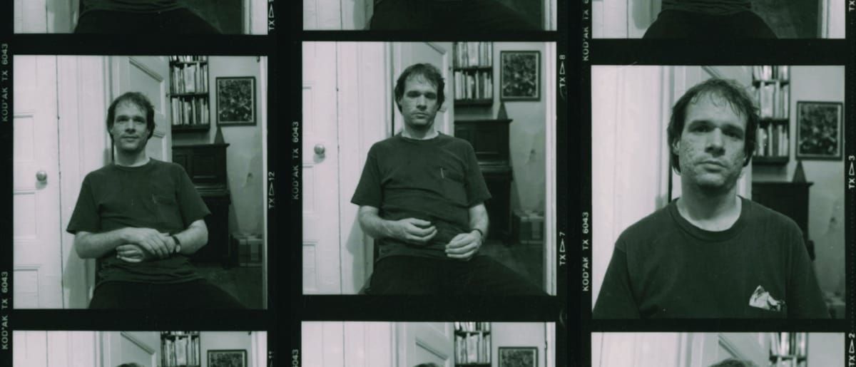 Contact sheet of black-and-white film frames showing Arthur Russell seated in a book-lined apartment, hands folded in his lap, in three slightly varied poses.