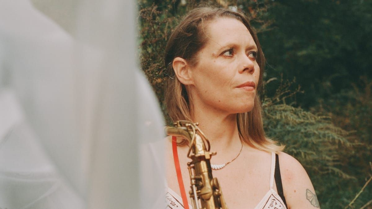Caroline Davis holds a saxophone at her side, gazing off-camera, standing outdoors before dense greenery. A blurred figure occupies the left foreground. Photo by Adi Meyerson.