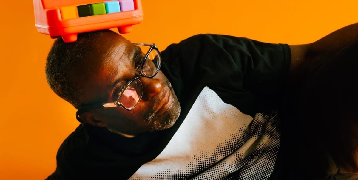 Charles Joseph Smith tilts his head toward the camera, balancing a colorful toy instrument on top of his head, wearing dark-framed glasses and a black jacket. Photo by Dennis Larance.