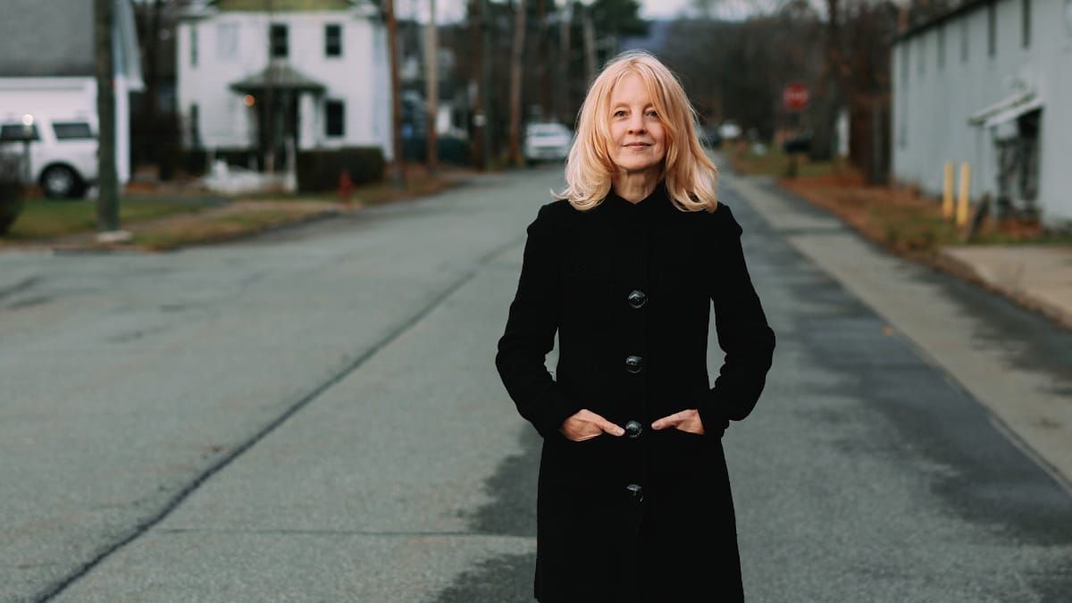 Maria Schneider stands in the middle of a quiet small-town street, hands in the pockets of a long black coat, a modest white clapboard building visible behind her. Photo by Briene Lermitte.
