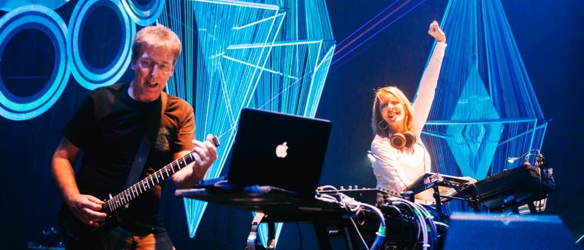 Steve Hillage plays electric guitar at left while Miquette Giraudy raises one arm behind a mixer and laptop, both performing against a vivid blue geometric light installation.