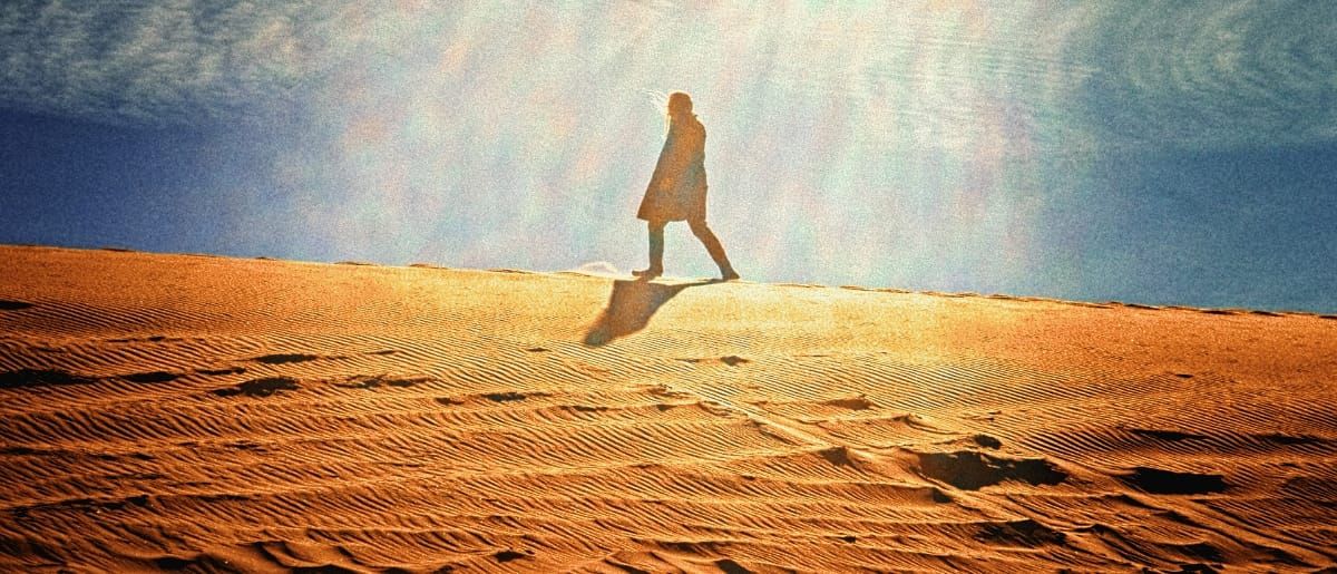 Johnathon Ford of Unwed Sailor walks along a sunlit sand dune, silhouetted against bright radiating light and a blue sky, long shadow trailing behind on rippled sand. Photo by Charles Elmore.