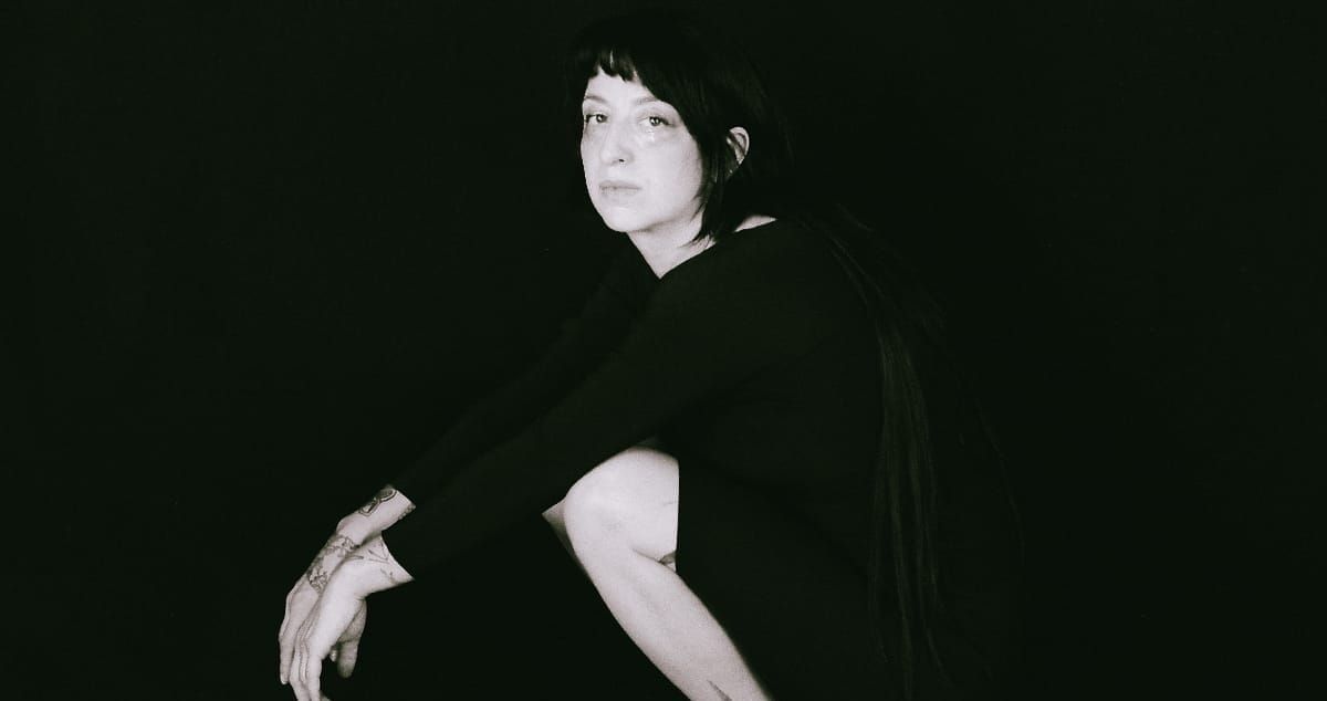 Ora Cogan stands against a black background in a black t-shirt, glancing over one shoulder, tattooed forearms crossed in front of her. Black and white.