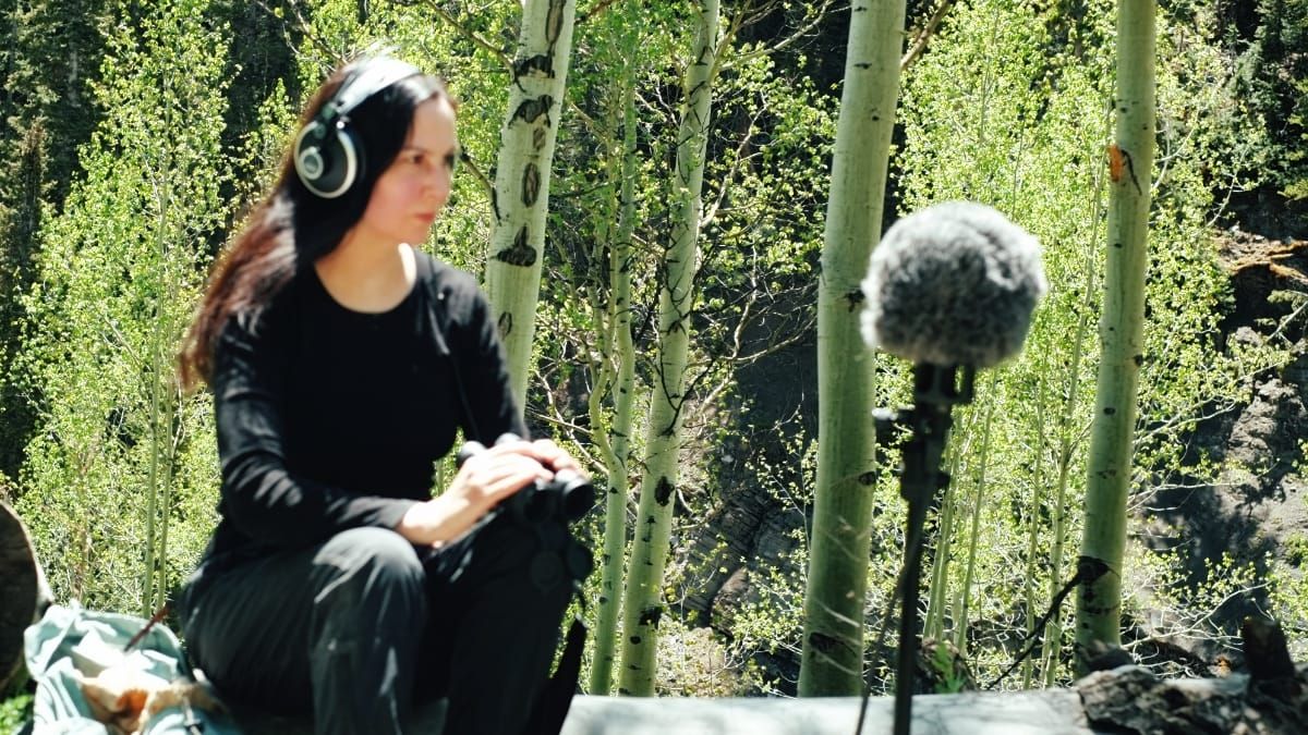 Patricia Wolf, dressed in black, crouches with headphones on, listening intently, while a boom microphone on a tripod faces them amid sunlit aspen trees.