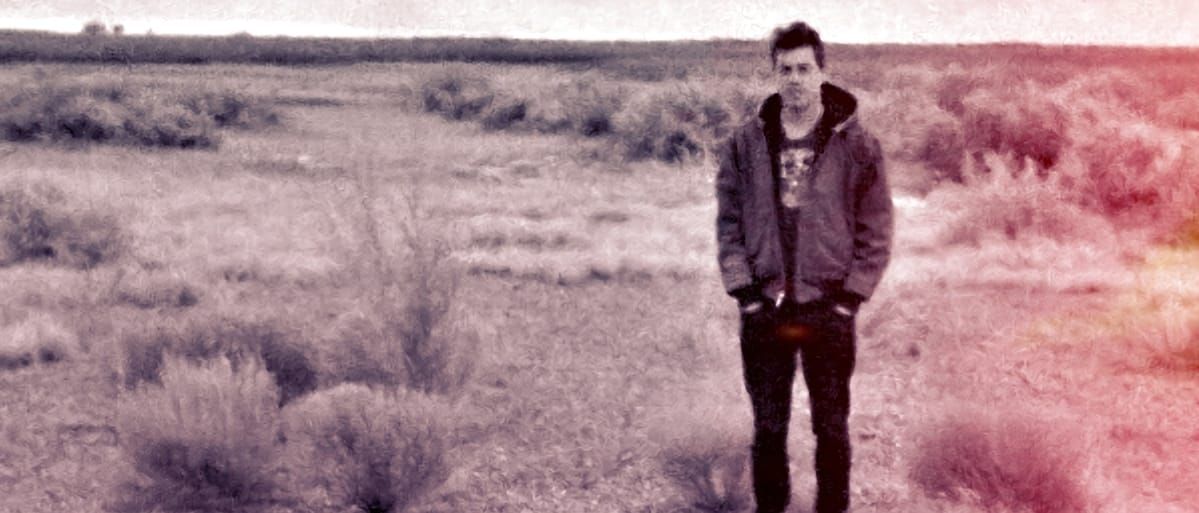 Phil Geraldi stands alone in an open desert scrubland, hands in pockets, wearing a hoodie and dark jeans. The photo has a faded quality with warm red-pink tones bleeding in from the right.