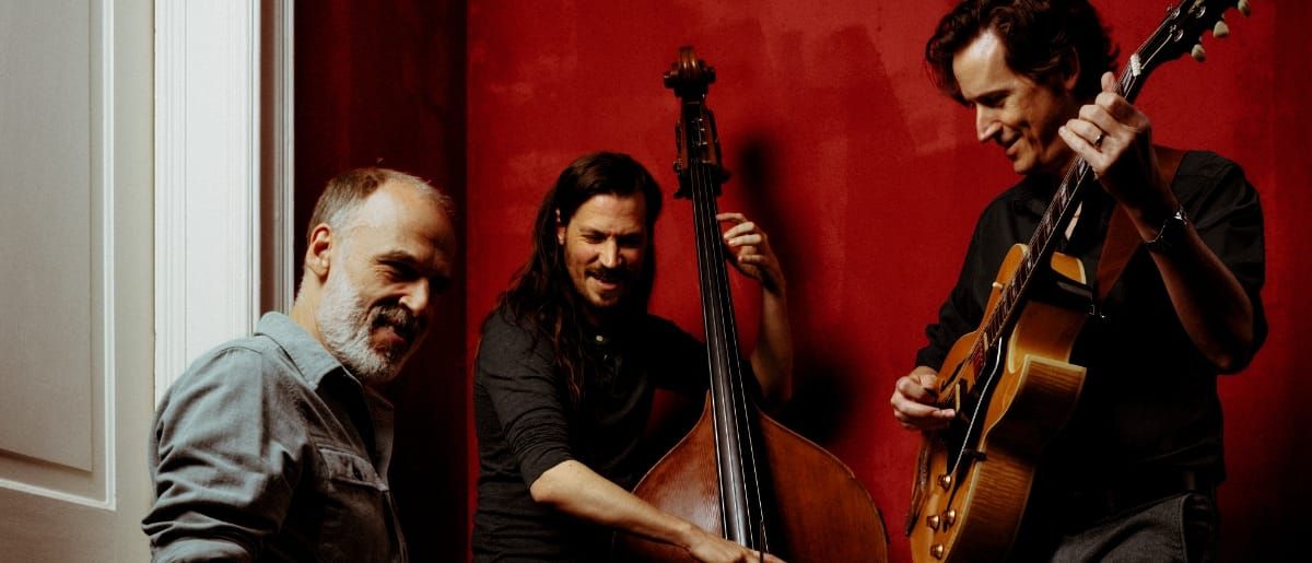 Three members of Cento Threeo against a deep red wall: one leans against a doorframe at left, one plays upright bass at center, one plays archtop guitar at right.