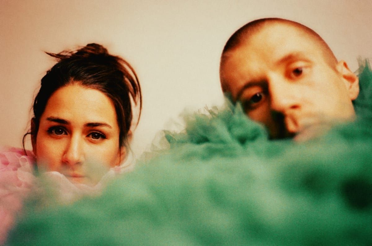 Curió Curió peer over a blurred haze of pink and green tulle, only their faces visible above the fabric, looking toward the camera.