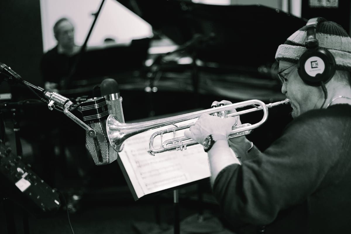 Dave Douglas plays trumpet in a recording studio, reading from sheet music on a stand, headphones on, a vintage microphone inches from the bell.