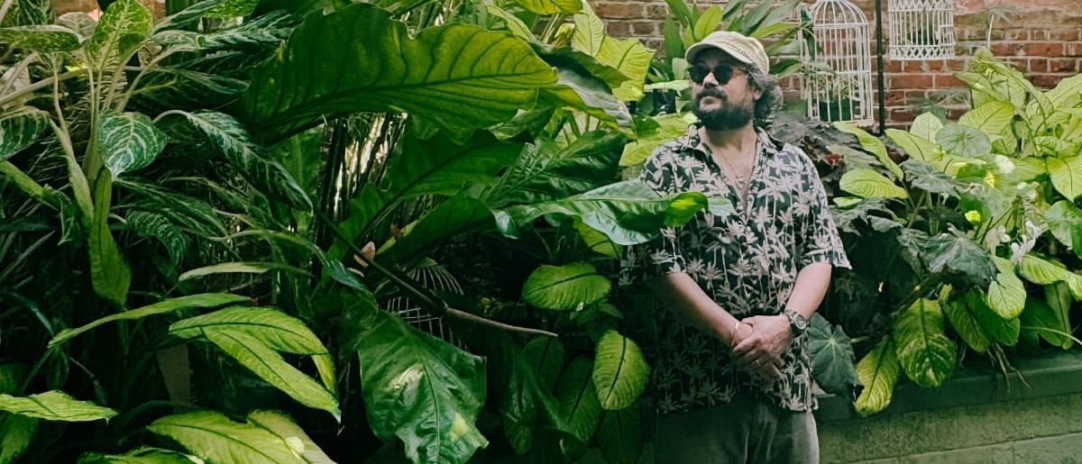M. Geddes Gengras stands among towering tropical plants with broad green leaves, wearing a cap, round sunglasses, and a patterned shirt, a brick wall and white birdcages behind him.