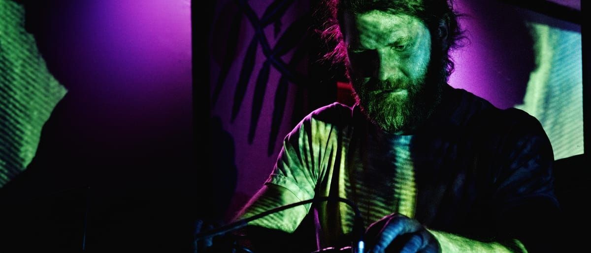Nolan Hildebrand, bearded with shoulder-length hair, leans over electronic gear mid-performance, lit in green and magenta stage light against a purple backdrop.