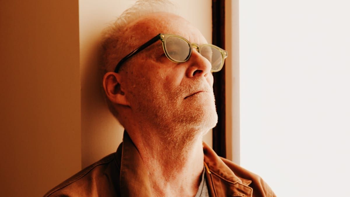Steven Hall leans his head back against a wall beside a bright window, chin lifted toward the light, wearing translucent green-framed glasses and a rust-colored jacket.