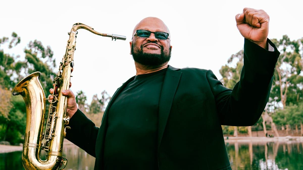 Azar Lawrence stands beside a lake holding a tenor saxophone in one hand, the other fist raised, in a black jacket and sunglasses.