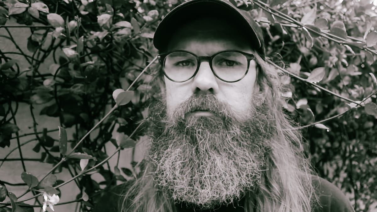 Josh Mason looks straight at the camera from behind leafy vines, face filling the frame, in a dark cap and round glasses, his long beard reaching the bottom edge. Black and white.