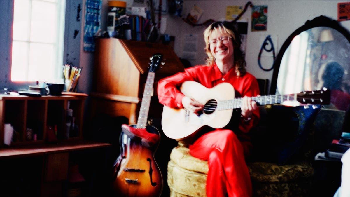 Sophie Wellington sits playing a pale acoustic guitar and laughing, dressed in red, in a warm cluttered room with an archtop guitar leaning beside her and light from a window.