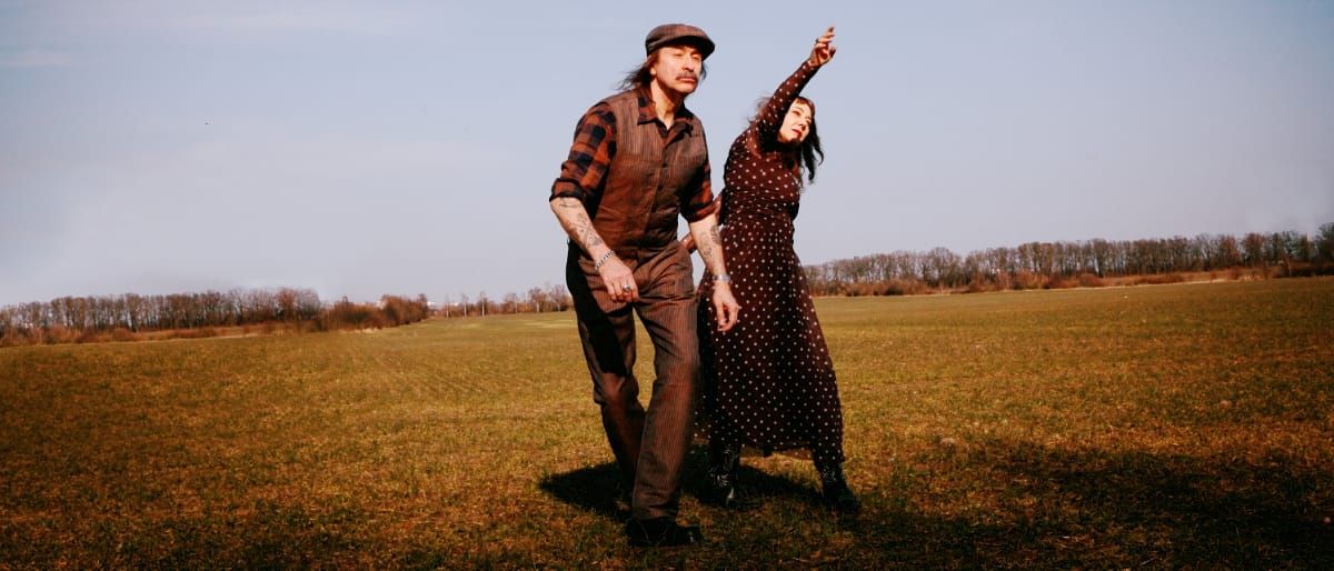 Alexander Hacke strides across an open field in a flat cap and pinstriped suit, one hand behind him linked to Danielle de Picciotto in a polka-dot dress, her arm flung skyward.