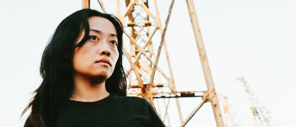 sachi's mirror stands below a rusted industrial crane, shot from a low angle against a pale sky, dark hair blown across one shoulder, looking off past the camera.