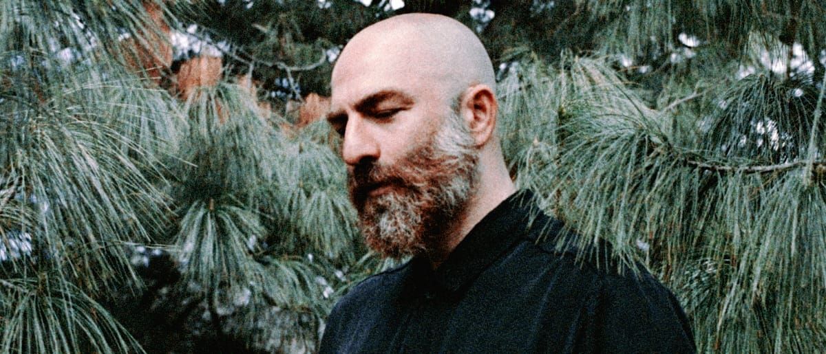 Deniz Cuylan looks down with his eyes closed, framed by the long needles of a pine, in a black shirt.