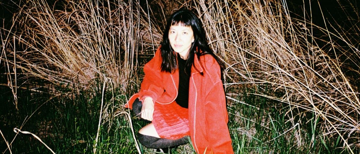 Frances Chang crouches among dry reeds and grass at night in a red coat and dark over-the-knee socks, looking into the camera.
