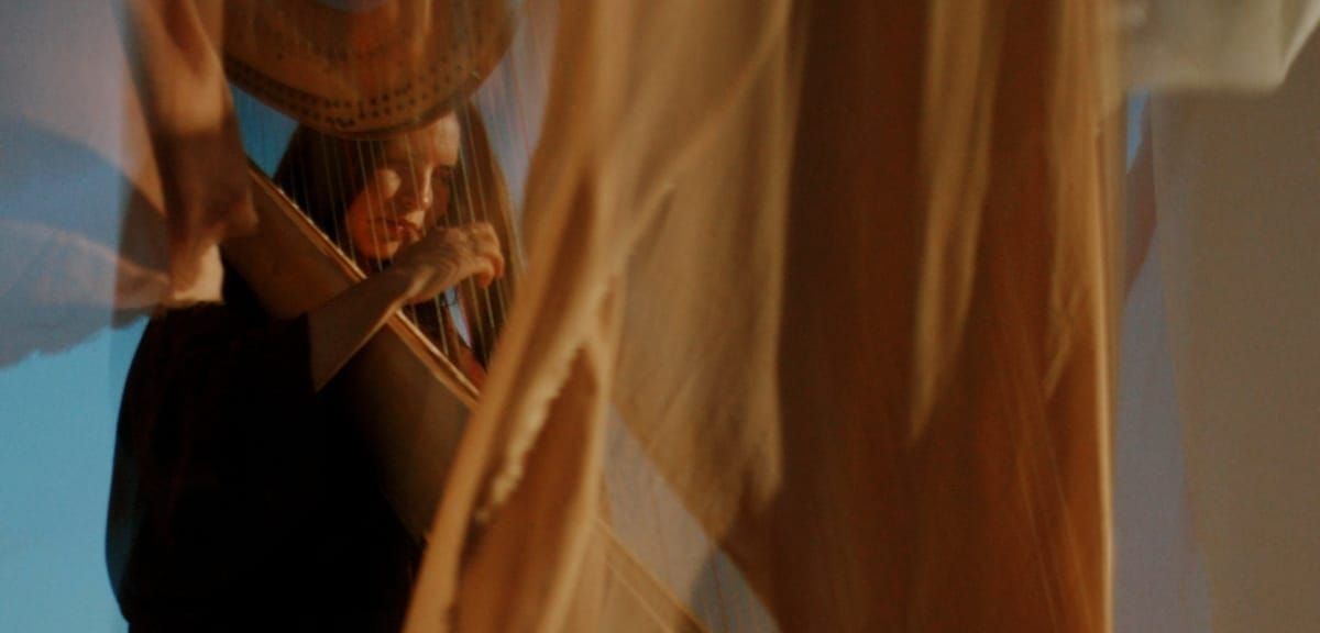 Lara Somogyi plays a harp behind drifting sheets of amber fabric, her face and hands glimpsed through the strings against a pale blue backdrop.