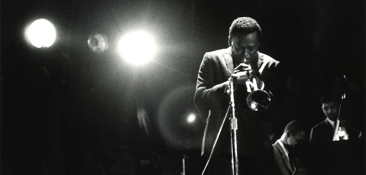 Miles Davis bends toward the mic under two bright stage lights, Wynton Kelly at the piano and Paul Chambers on bass behind him in the dark.