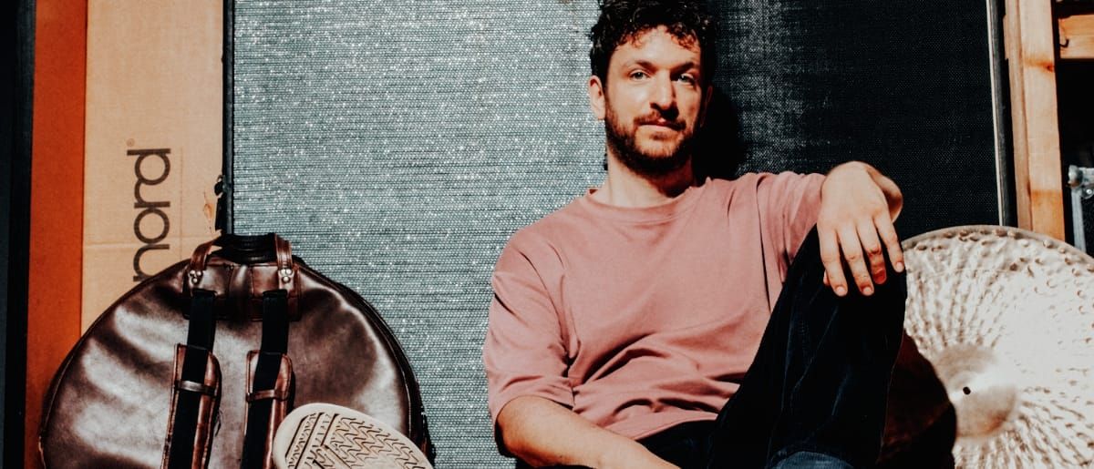 Peter Gall sits on the floor against a textured silver panel in a pink T-shirt, one knee up, a leather cymbal bag and an orange Nord box to his left and a large cymbal to his right.