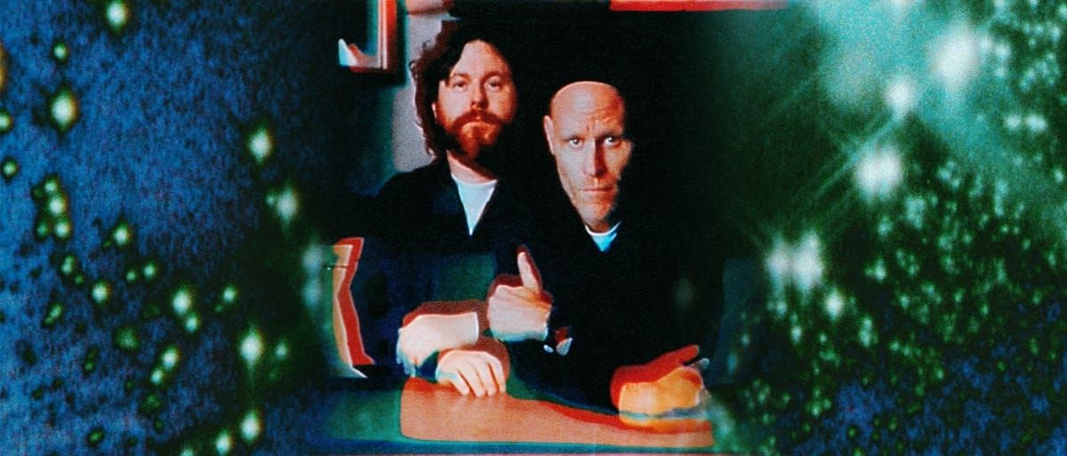 Gareth Quinn Redmond and Francis Harris sit together at a table in a grainy, color-shifted film still, framed by speckled blue and green light.