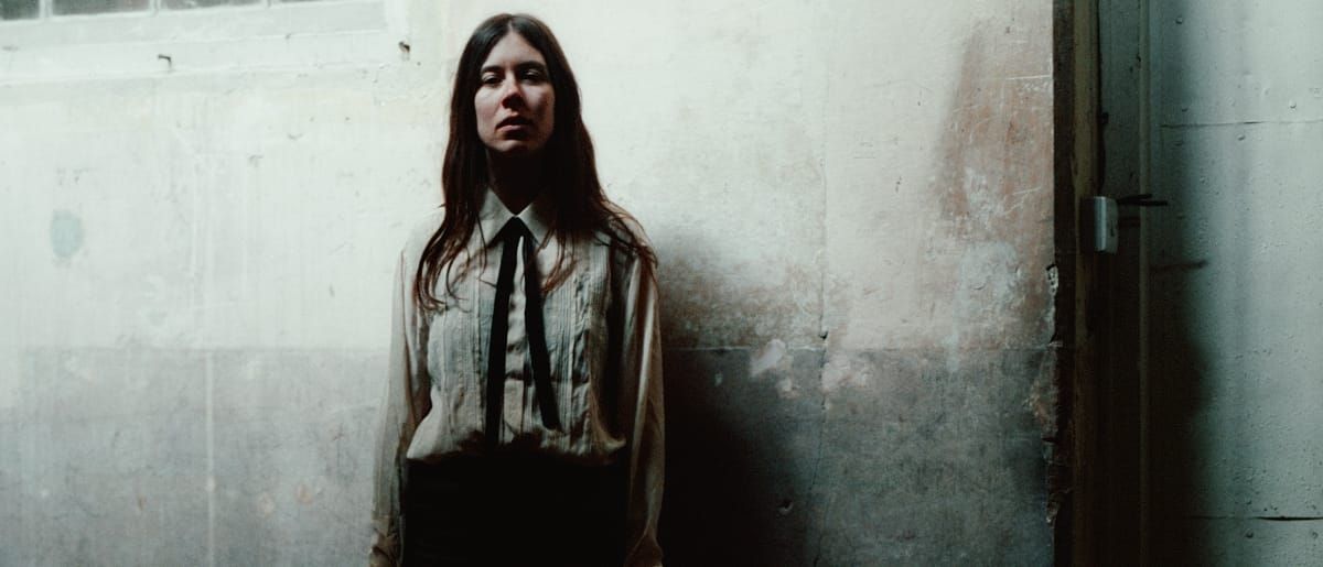 Nina Winder-Lind stands against a stained plaster wall in a pleated white shirt and thin black tie, her shadow beside her, gazing at the camera.