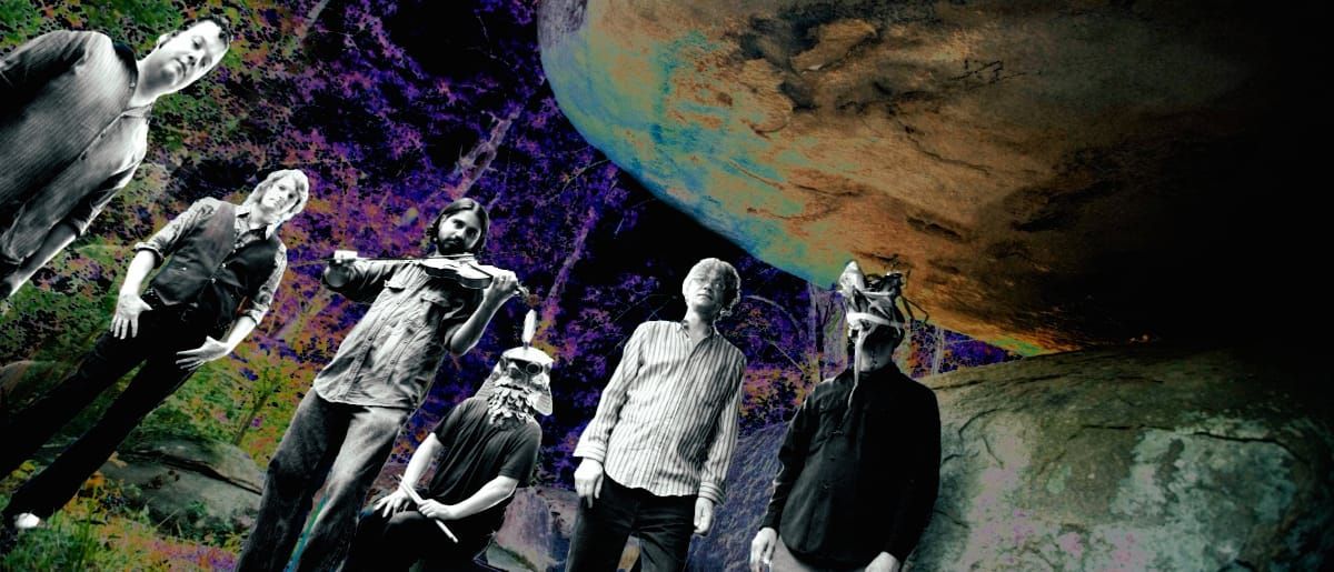 The Olivia Tremor Control, six members seen from below beneath a huge overhanging boulder in a purple-tinted forest; one holds a violin, two wear bird-like masks.