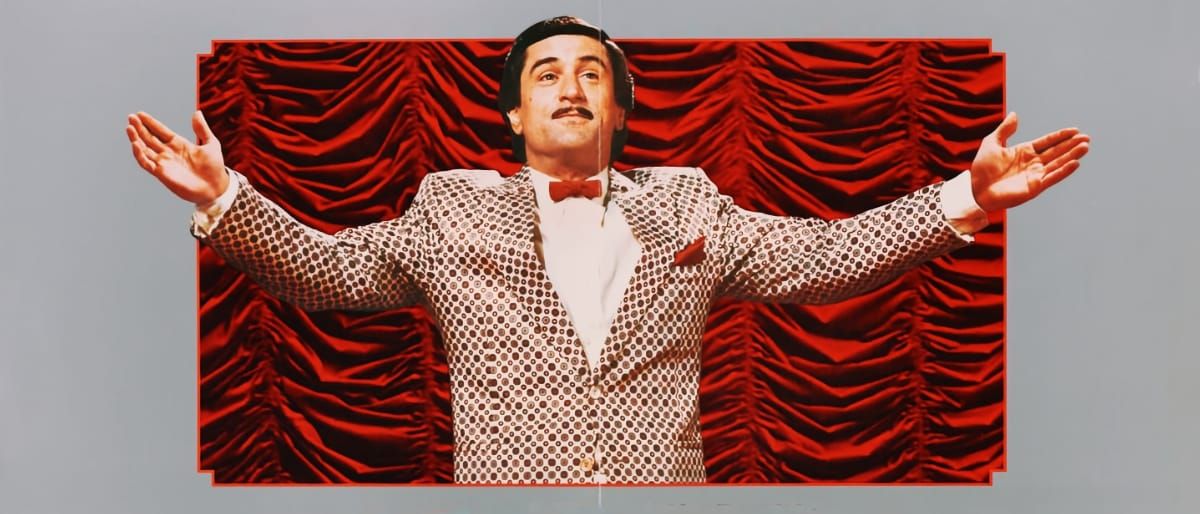 Robert De Niro as Rupert Pupkin stands arms outstretched in a sequined jacket and bow tie before red stage curtains, in a promotional image from The King of Comedy.