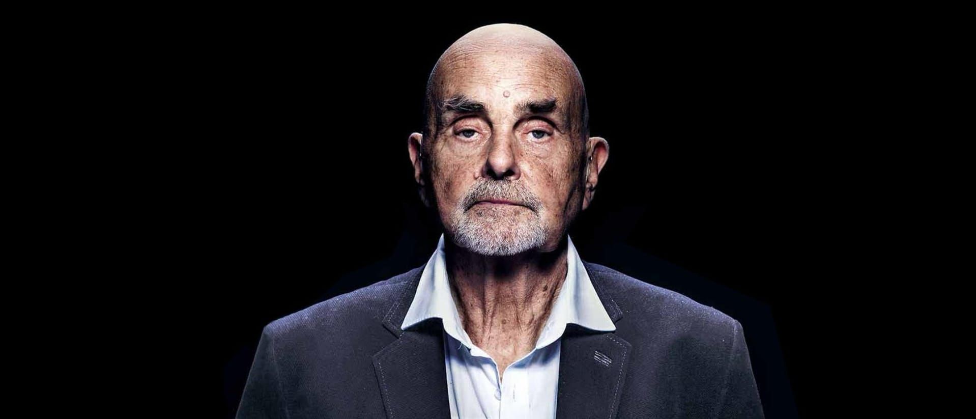 A stark studio portrait of Hans-Joachim Roedelius, bald and white-bearded in a grey blazer and open-collar shirt, staring directly into the camera against a pure black background.