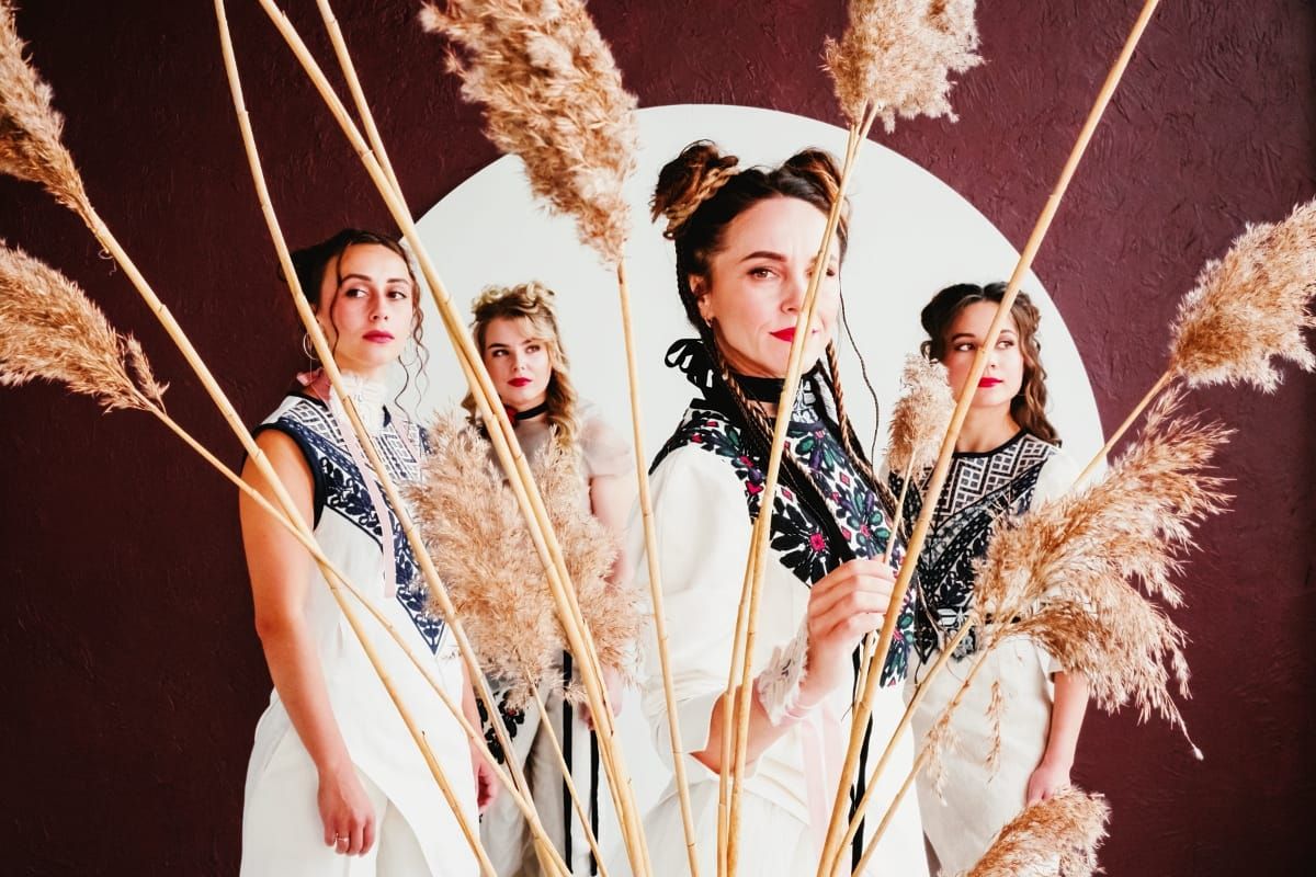 Four members of YAGÓDY in white embroidered traditional garments stand amid tall dried pampas grass stems before a circular white form on a deep burgundy background.
