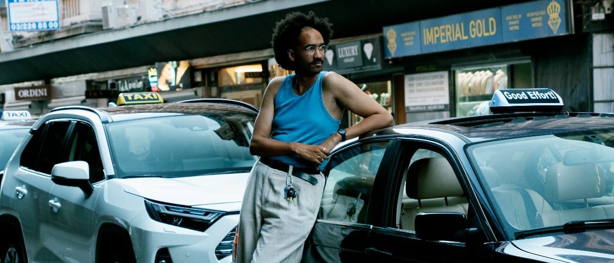 Youniss leans against a taxi on a busy city street in a blue tank top and grey trousers, with shop fronts and a second cab reading "Good Effort!" visible behind him. Photo by Adel Setta.