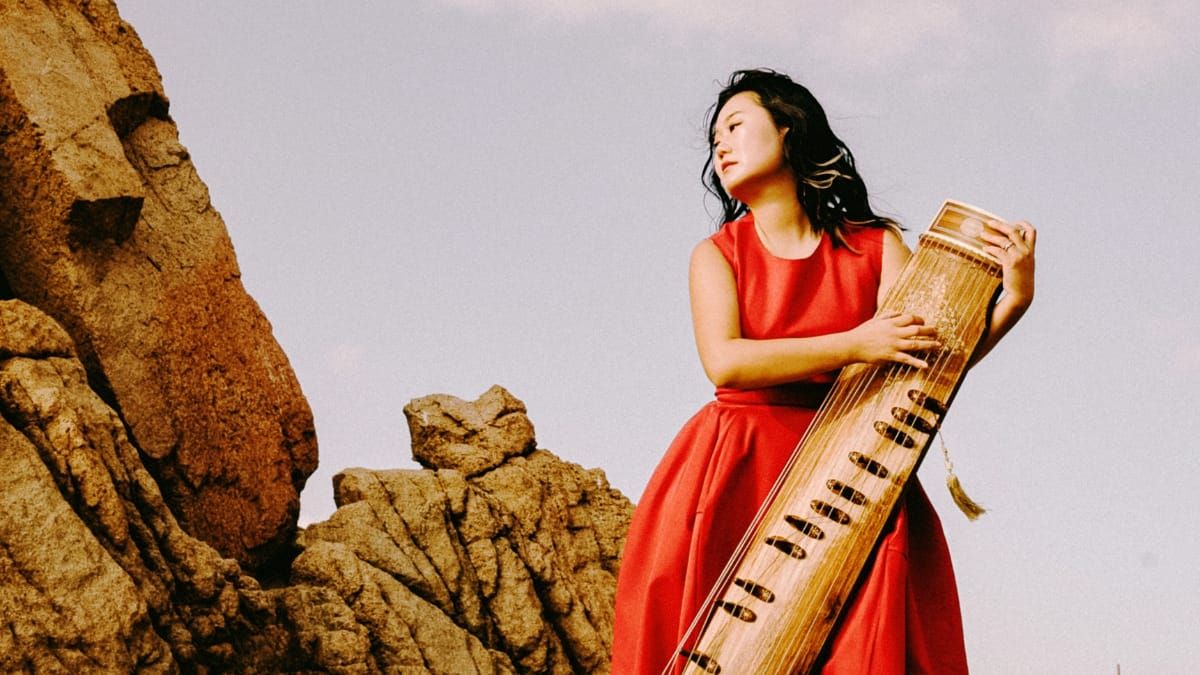 DoYeon Kim stands among desert rocks in a red dress, holding a gayageum against a pale blue sky.