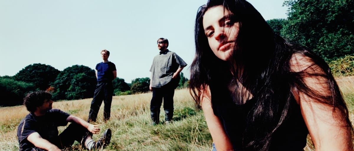 One member of Modern Woman leans close to the camera, long dark hair windswept across her face, while three others stand and sit in a sun-bleached field behind them.