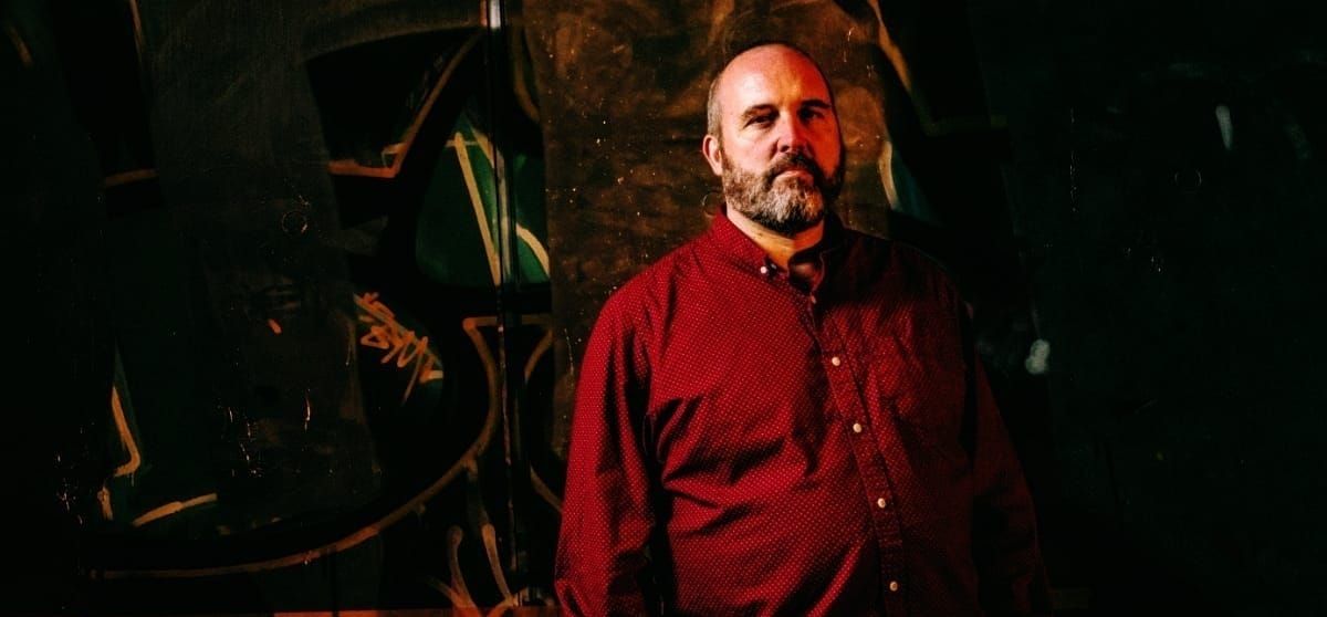 Nick Fraser stands against a dark wall covered in graffiti, wearing a deep red button-down shirt, arms at his sides, with a full beard and a steady, neutral gaze toward the camera.