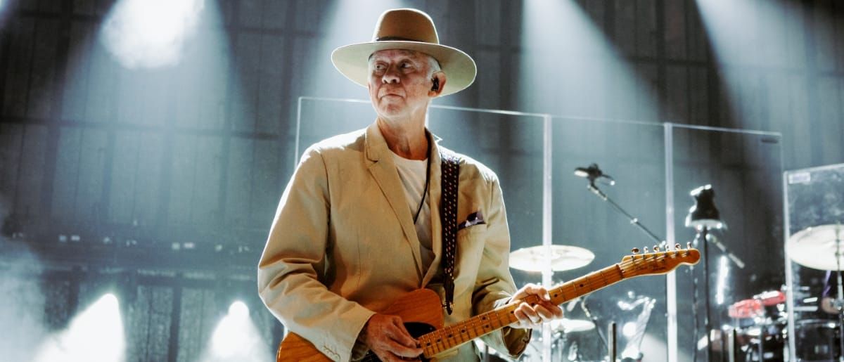 Phil Manzanera performs onstage holding a Fender Telecaster, wearing a cream suit and wide-brimmed hat, lit by dramatic stage lighting against an arched industrial backdrop.