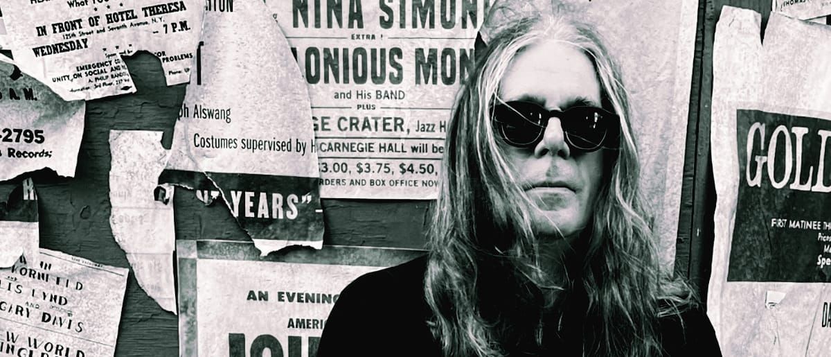 Bob Bert in dark sunglasses before a wall of torn vintage concert posters featuring Nina Simone and Thelonious Monk. Black-and-white. Credit: Michael Jung.