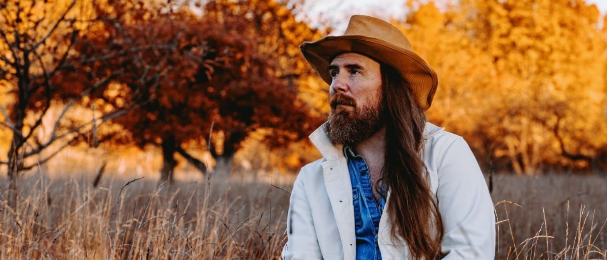 Bob Wagner stands in a dry autumn field, long hair and beard, wearing a tan wide-brim hat and white jacket over a denim shirt, gazing to one side against a backdrop of amber-lit trees.
