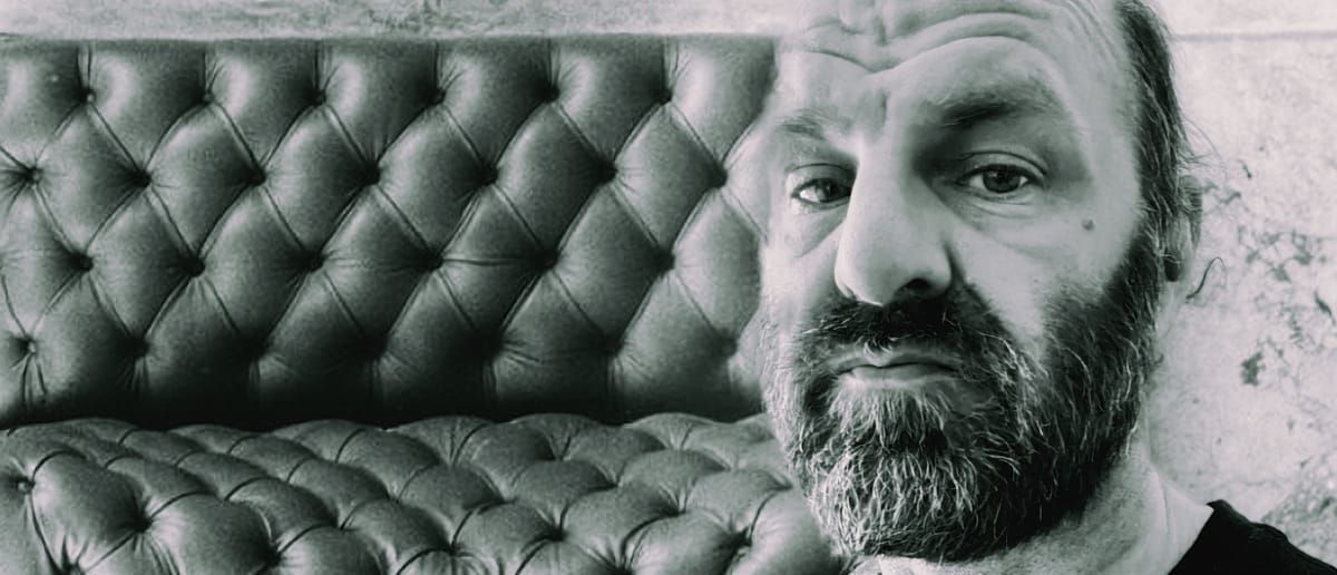 Chik White, bearded, gazes into the camera in a black-and-white close-up, seated against a tufted leather couch with a cracked plaster wall behind him.