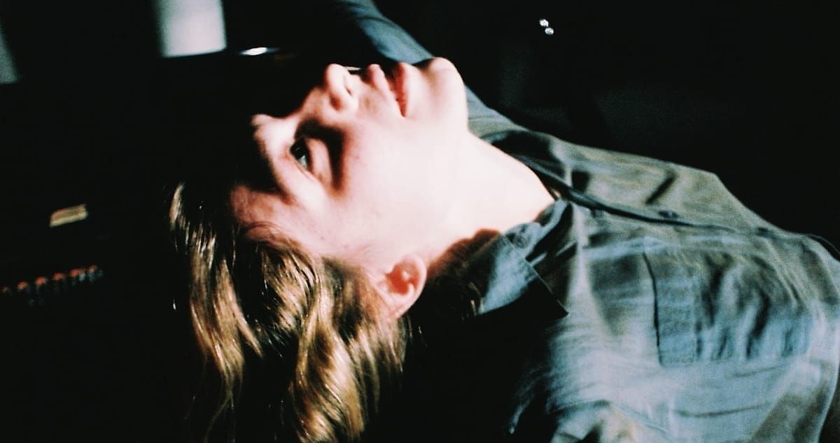 Eliana Glass reclines with head tilted back and eyes closed, shot on grainy film against a dark background, a piano visible at the left edge of the frame.