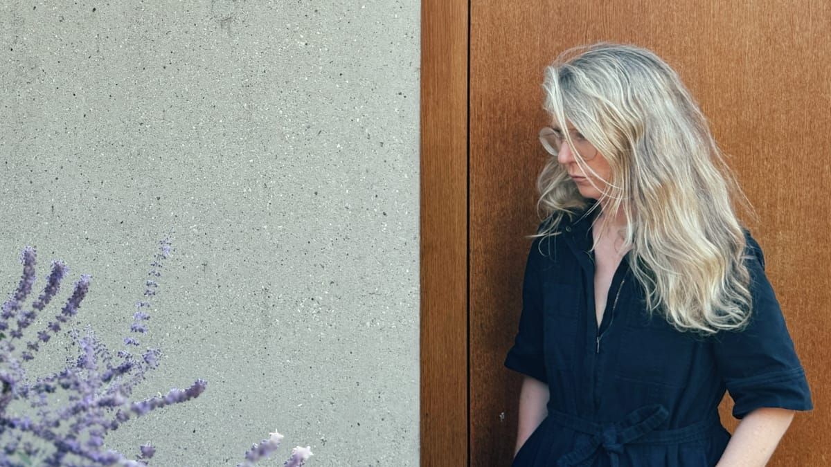 Félicia Atkinson stands beside a concrete wall and wooden door, long silver hair blown across her face, wearing a dark jumpsuit. Lavender blooms in the foreground.
