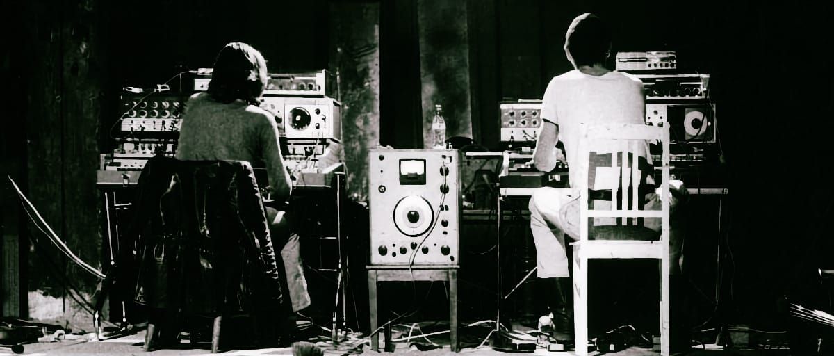 Black-and-white photo: two performers sit with backs to camera, each facing a rack of vintage electronic equipment. A large unit with a VU meter stands between them on a bare stage.