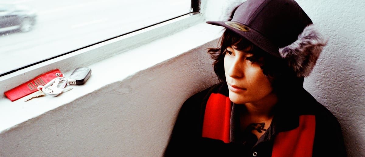 zzzahara seated against a wall beneath a window, wearing a fur-eared cap and red-and-black jacket, gazing upward. Keys and earbuds rest on the sill behind them. Photo by Lindsey Byrnes.