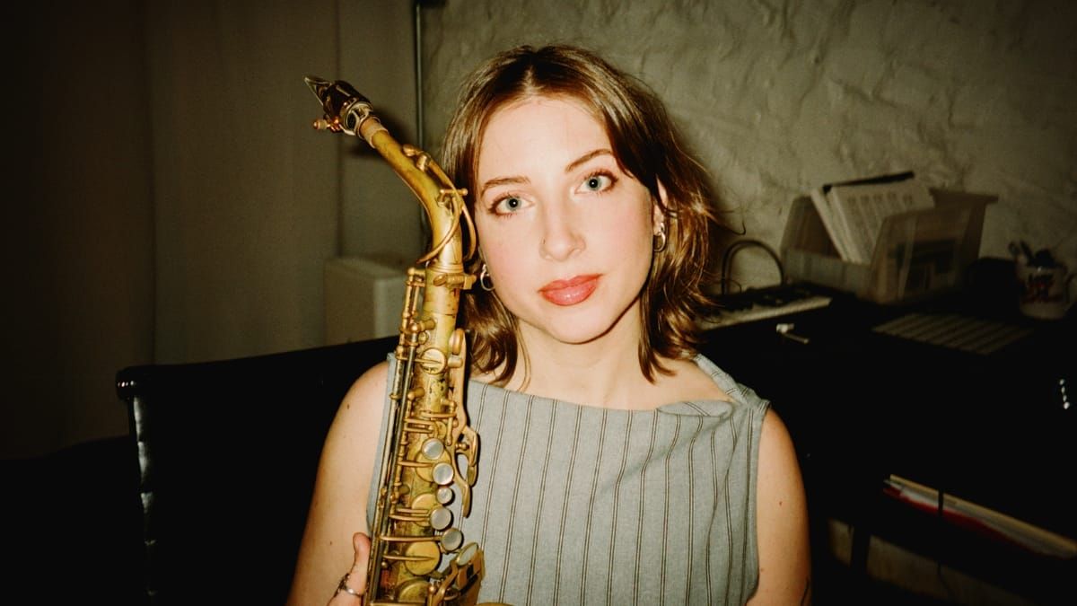 Alden Hellmuth holds an alto saxophone upright against her cheek, looking at the camera in a dim room with a keyboard and sheet music visible behind her.