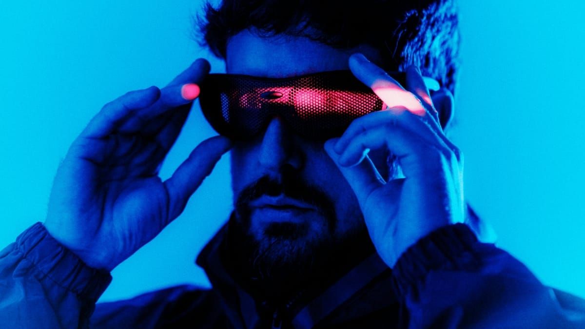 Danalogue holds a pair of red-lit mesh visor glasses to his face with both hands, his bearded face and jacket washed in deep blue against a bright blue background.