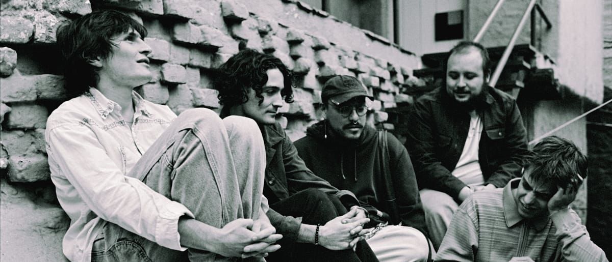 Diles que no me maten sit together against a brick wall in black and white, five members leaning and talking, one resting his head on his hand.